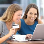 Two happy friends paying on line with a credit card and a laptop sitting in a bar terrace