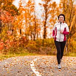 Middle-age Asian woman running on a park path through autumn foliage.