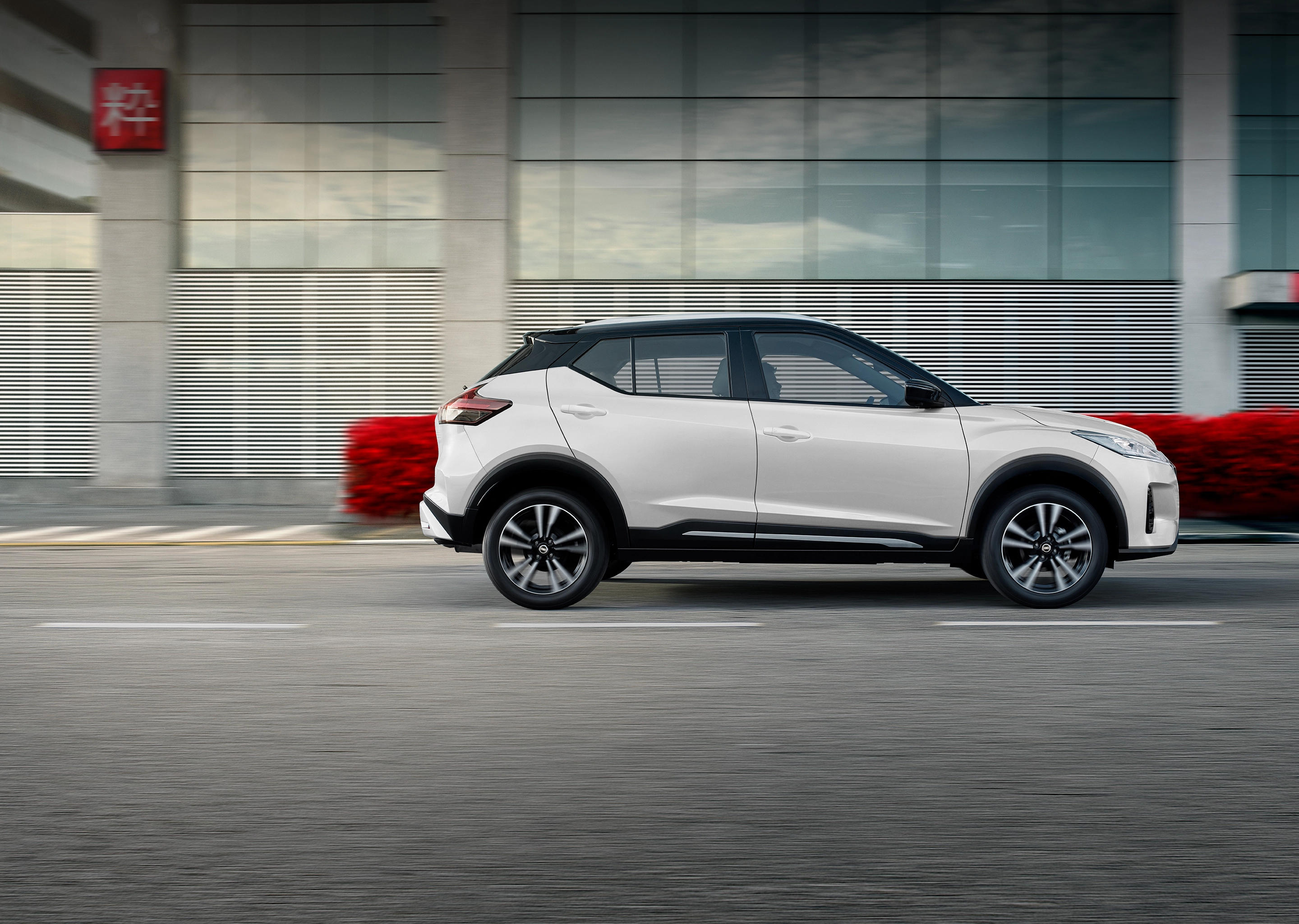 Nissan Kicks branco em movimento na estrada, com um fundo de edifício moderno e vegetação ao lado.