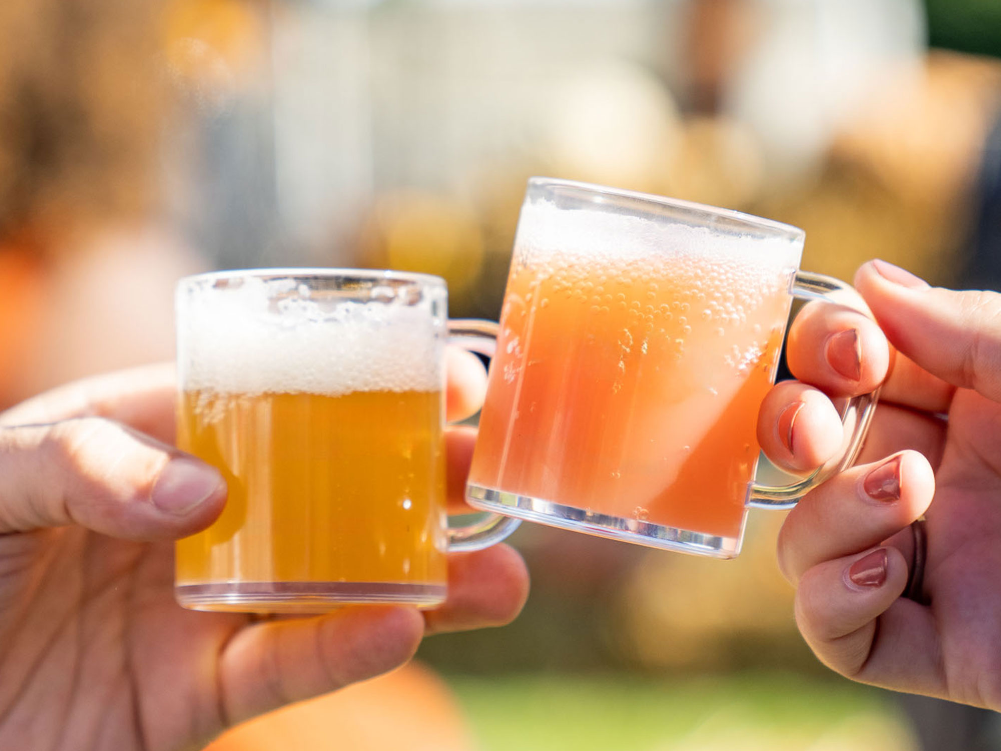 Two people clink sample mugs of beer outdoors on a sunny day