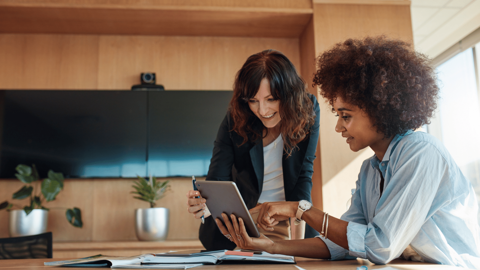 A black woman, sitting down, is looking over a tablet with her coworker, a white woman, as she stands hunched over. Both women are smiling 