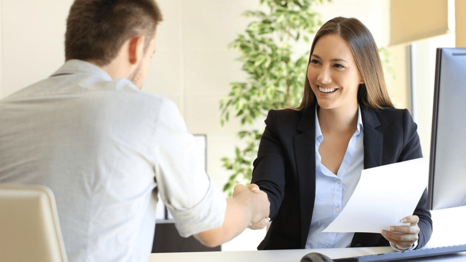 woman sitting at desk, holding a piece of paper, and shaking hands with a man across from her desk