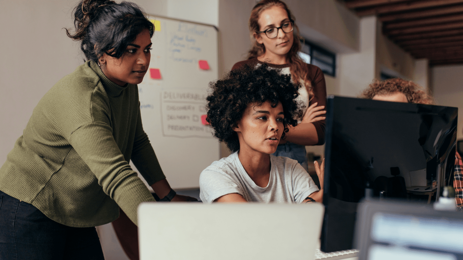 Three women and a fourth person (whose head you can only see part of) surround a computer, working on something. in the background, there's a white board with the word 