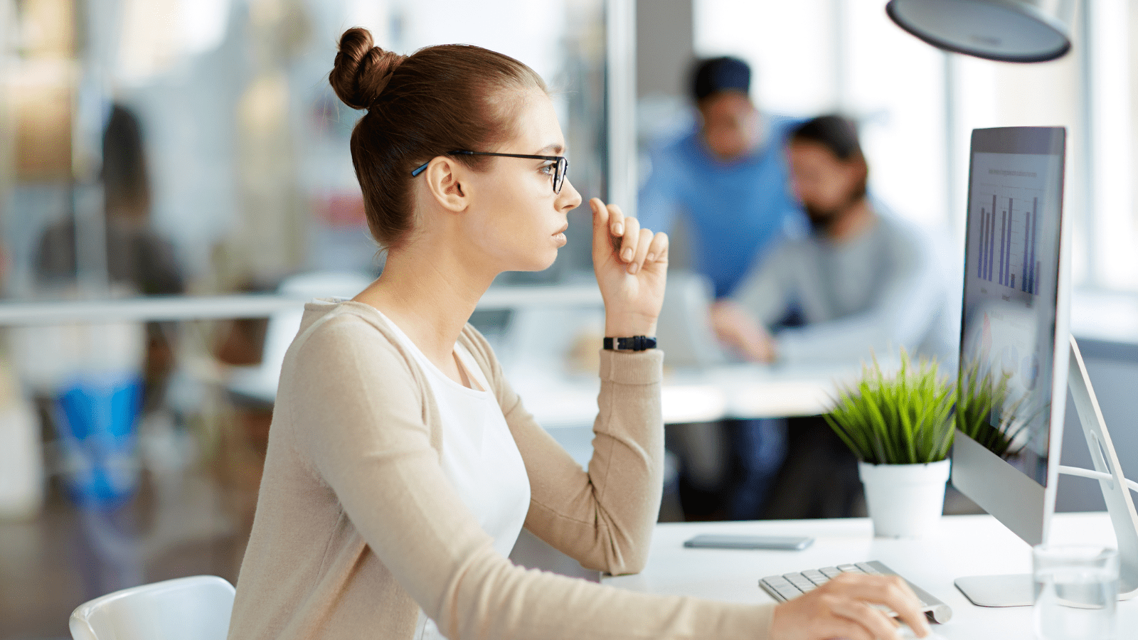 woman is sitting at her computer, looking at a chart. Two men are in the background looking at another computer.