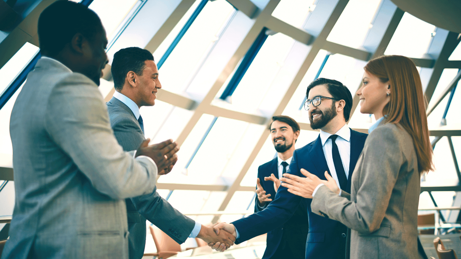 A group of business people, three on one side and two on the other, are standing together and smiling. One from either side are shaking hands. 