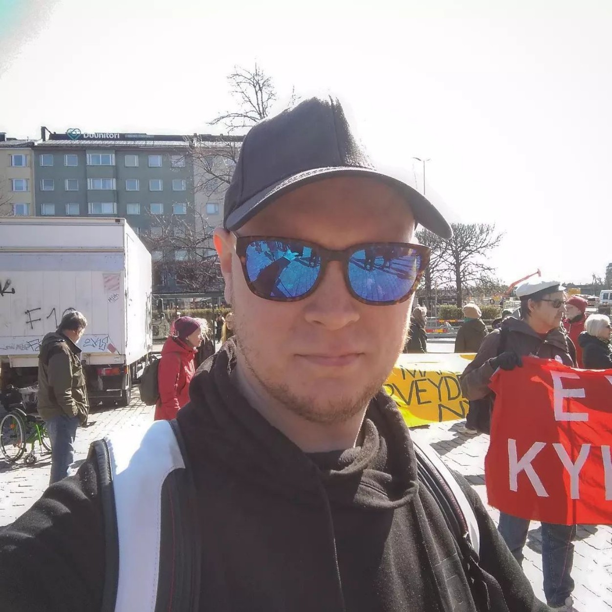 Selfie of Otto Rask on a sunny day, taking part in a political rally. He's wearing a cap and blue-tinted sunglasses.