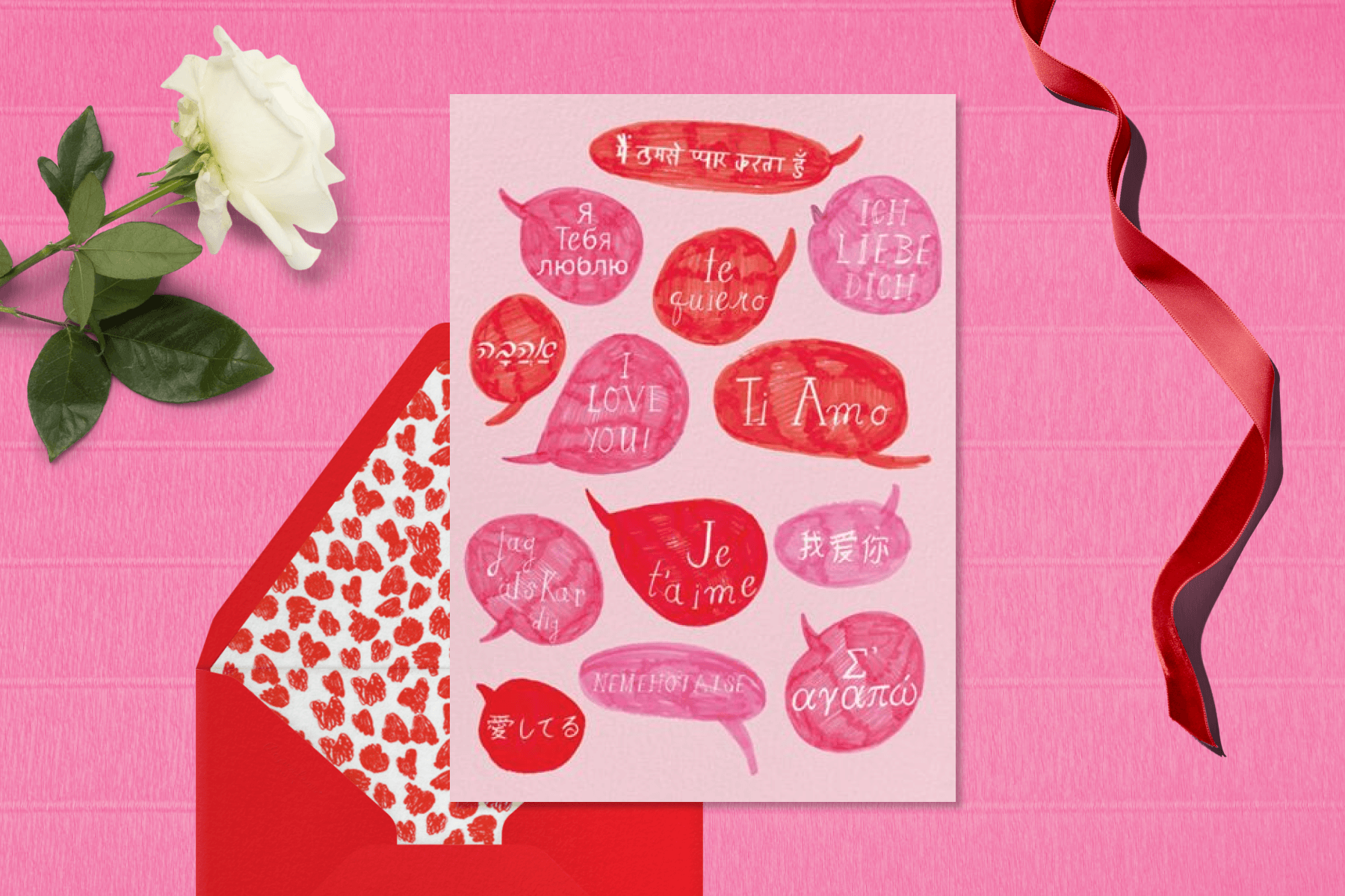 A pink Valentine's Day card with text bubbles filled with the phrase "I love you" in different languages, paired with a red envelope with red and white hearts liner. The card is surrounded by a white rose and a red ribbon.