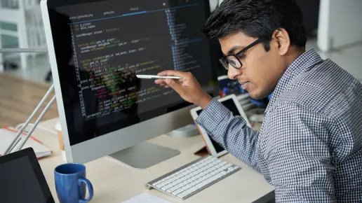 Image of a student in front of a computer, pointing at the screen with a writing pen while looking down at a notebook.