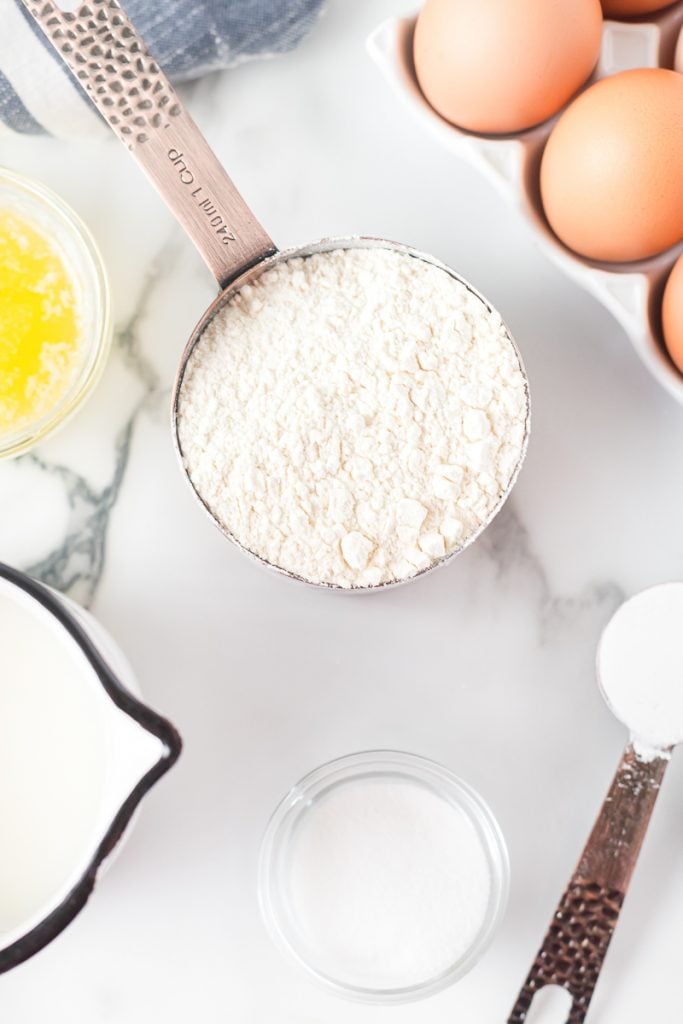 flour, butter, eggs in measuring cups laid out to make pancakes. 