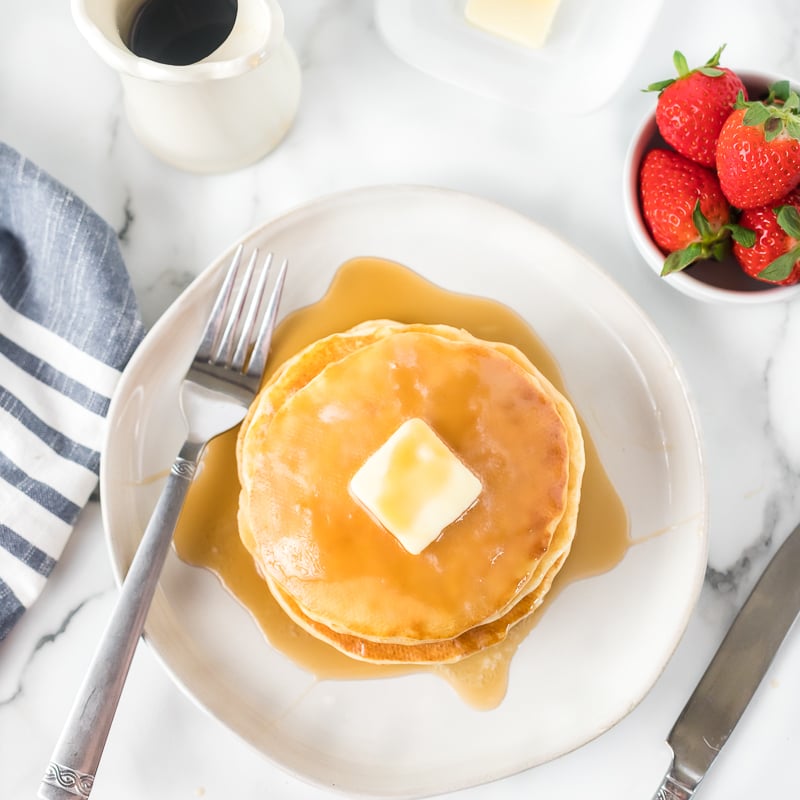 overhead shot of pancakes with butter & syrup with a side of strawberries.