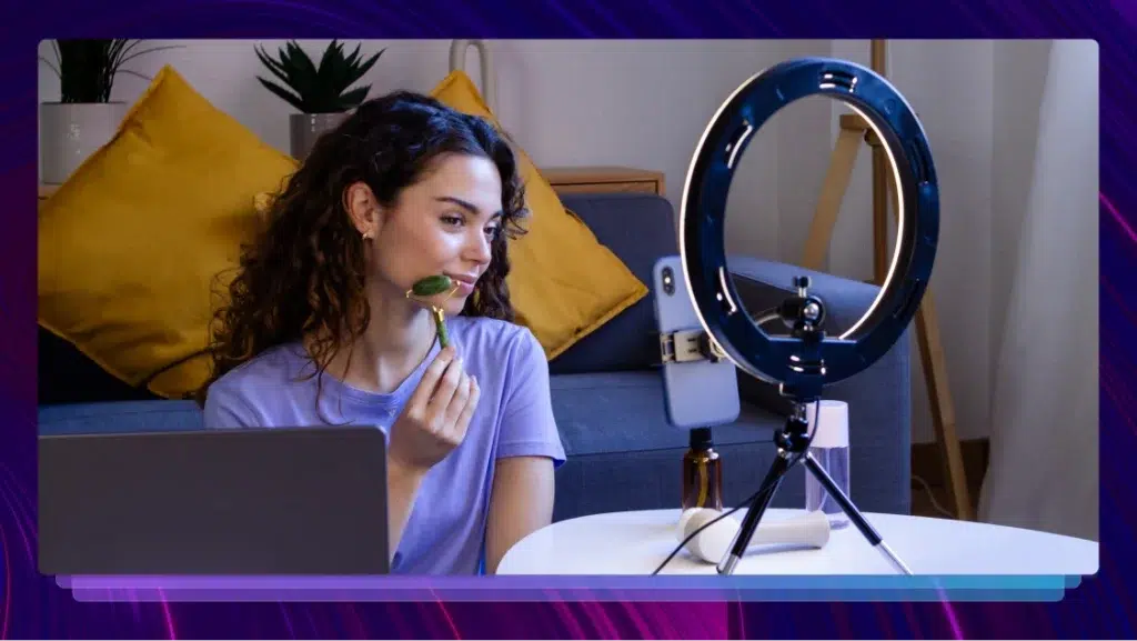 A woman records herself with a cellphone and a ring light as she uses a jade roller. She has a laptop and other beauty products next to her.