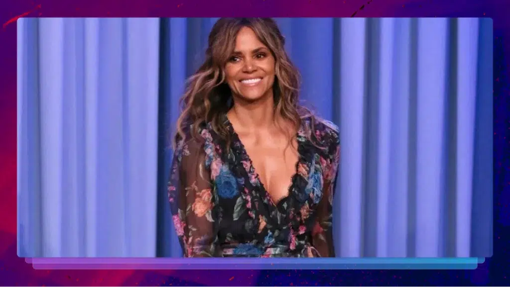 Halle Berry smiles and stands in front of a purple curtain, with a purple border around the image.