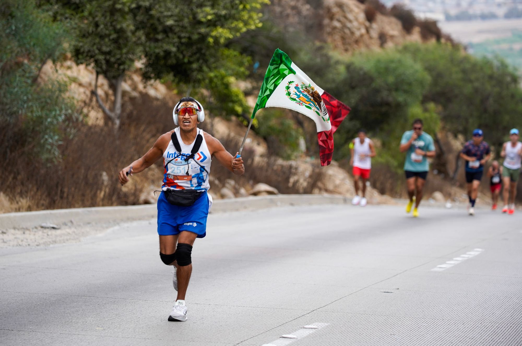 Runners compete in the Binational Race, from Las Americas Premium Outlets in San Ysidro to Playas de Tijuana. A runners carrying a Mexico flag on the Via International hill along the US/Mexico Border in Colonia Soler on Sunday, Sept. 15, 2024 in Tijuana, Baja California. (Alejandro Tamayo / The San Diego Union-Tribune)