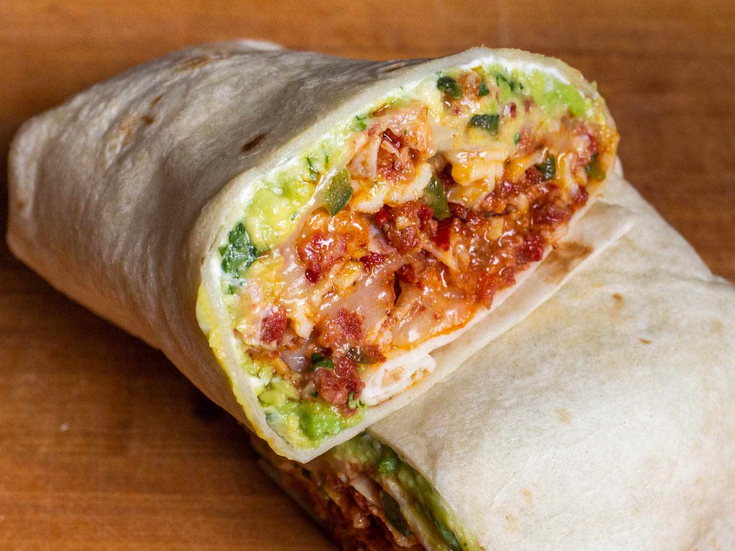 A breakfast burrito cut in half to show filling: creamy refried beans, scrambled eggs with chorizo, guacamole, crema (or sour cream), and hot sauce. It looks high-moisture in the best way, not wet or soggy.