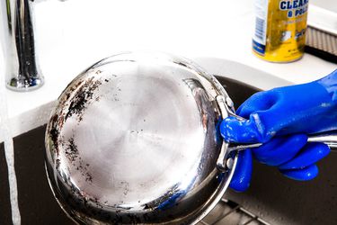 A blue-gloved hand holding a stainless steel skillet under a faucet