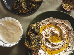 Overhead view of a plate of labneh with za'atar pita chips surrounded by a grocery store container of labneh, a bowl of zatar, and a plate of pita chips