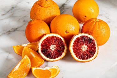 Whole and cut up oranges against a white marble countertop.