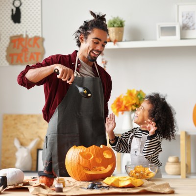 Smiling african american parent father removing pulp from ripe pumpkin while carving jack o lantern with little son for Halloween celebration at home in kitchen and looking at each other with smile