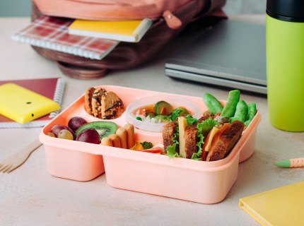 Healthy food concept: Open lunch box with sandwiches, vegetable salad, fresh grape and kiwi, rice and nuts on the table near backpack, laptop and thermo mug; selective focus