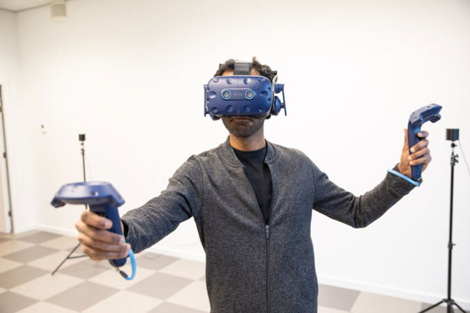 A man wearing a black undershirt and a gray jacket is wearing a blue VR headset that covers most of his face over his head. His arms are out, and each hand is holding a blue controller. He is standing in a room with white walls and checkered floor, and there are two tripods standing in the background.