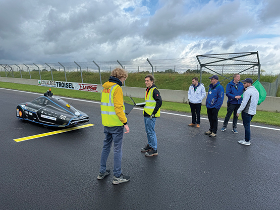 With the black vehicle in the background, two people in yellow vests talk with laptops in their hands. Team members mingle in the right corner of the photo. 