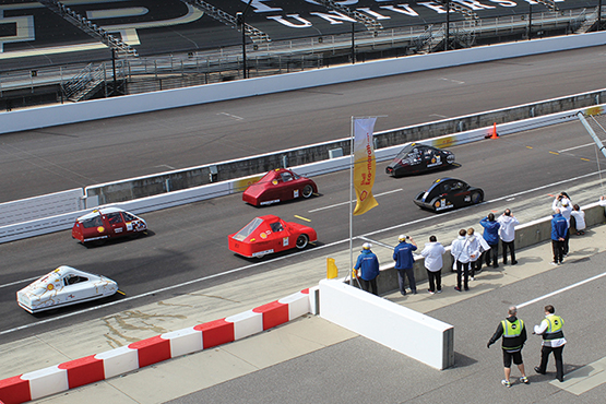 Six vehicles on the racetrack, three red, two black, one white. A group of people watch them from the sidelines.