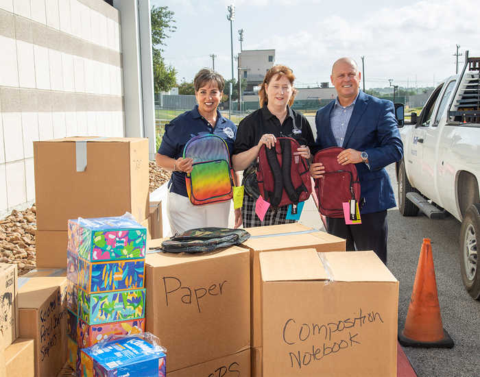 two women and 1 man holding backpacks standing behind cardboard boxes labeled with types of school supplies 