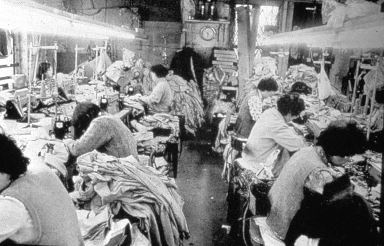 Garment factory in Chinatown where seven adults are working at sewing machine stations with overflowing piles of fabric