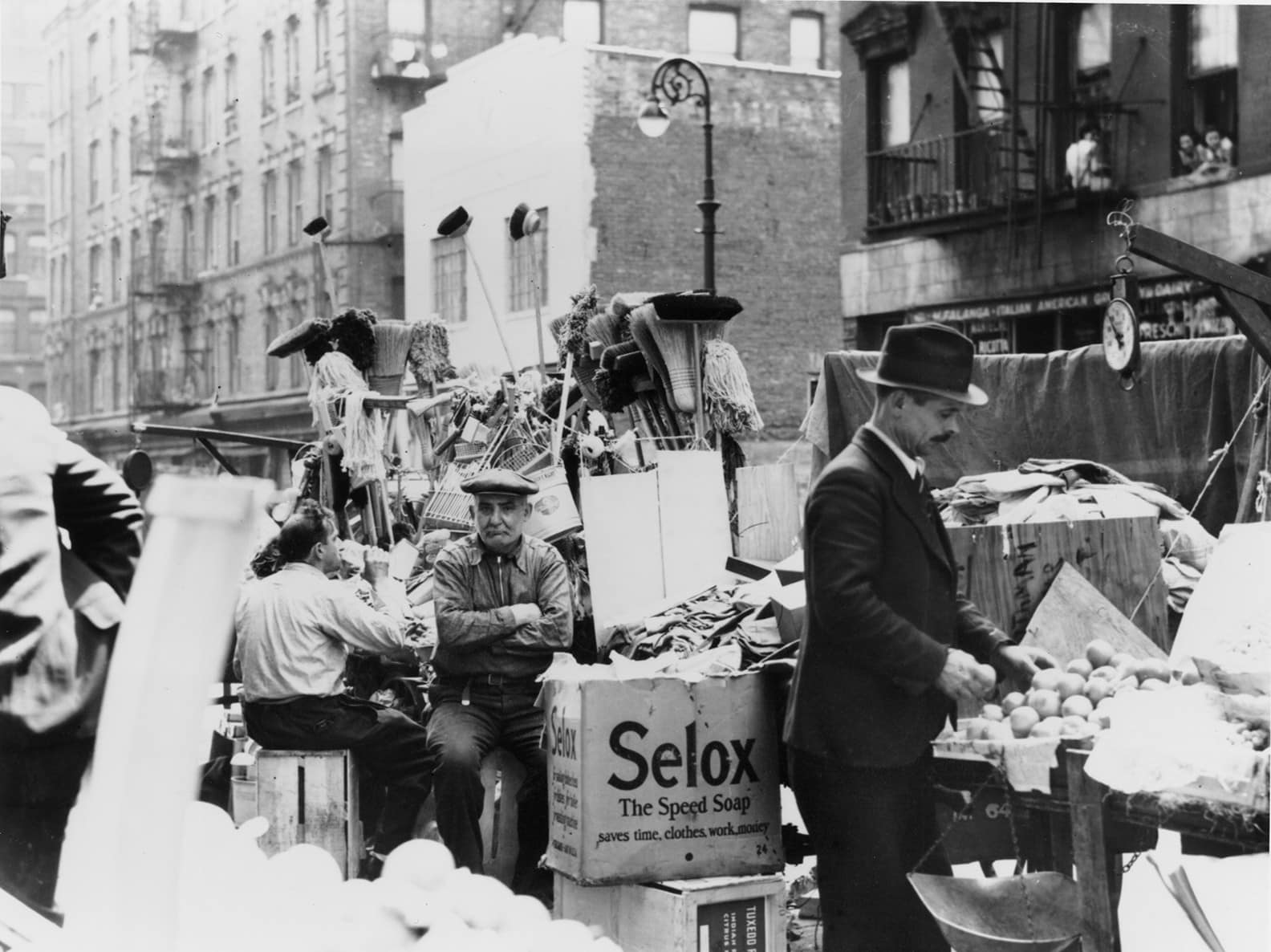 A couple of pushcarts surround a shopkeeper who is frowning while sitting on a crate with the brooms and mops they're selling piled behind them