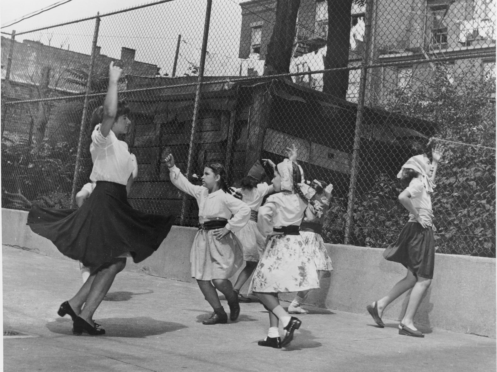 An adult with light skin tone teaching 6 young children in skirts and braids how to dance in a fenced concrete yard