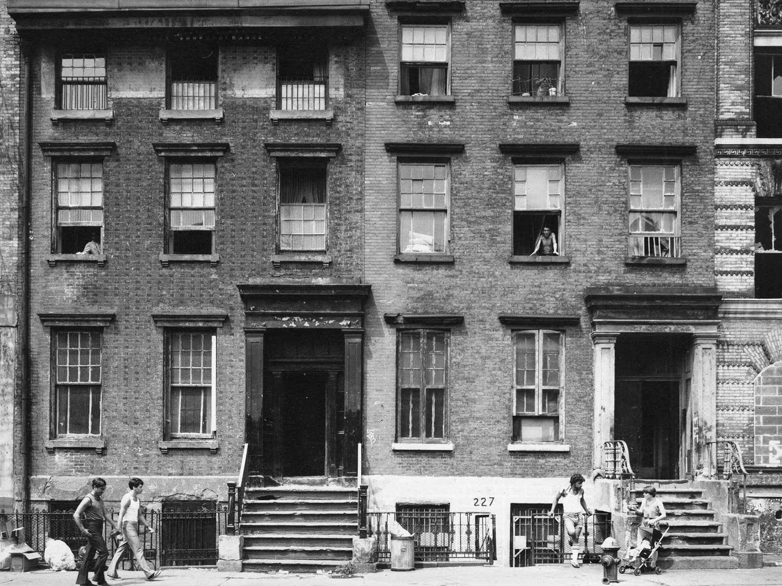 Photo of tenements with people walking by on the sidewalk, looking out from their windows, and lounging on the building steps in the summer