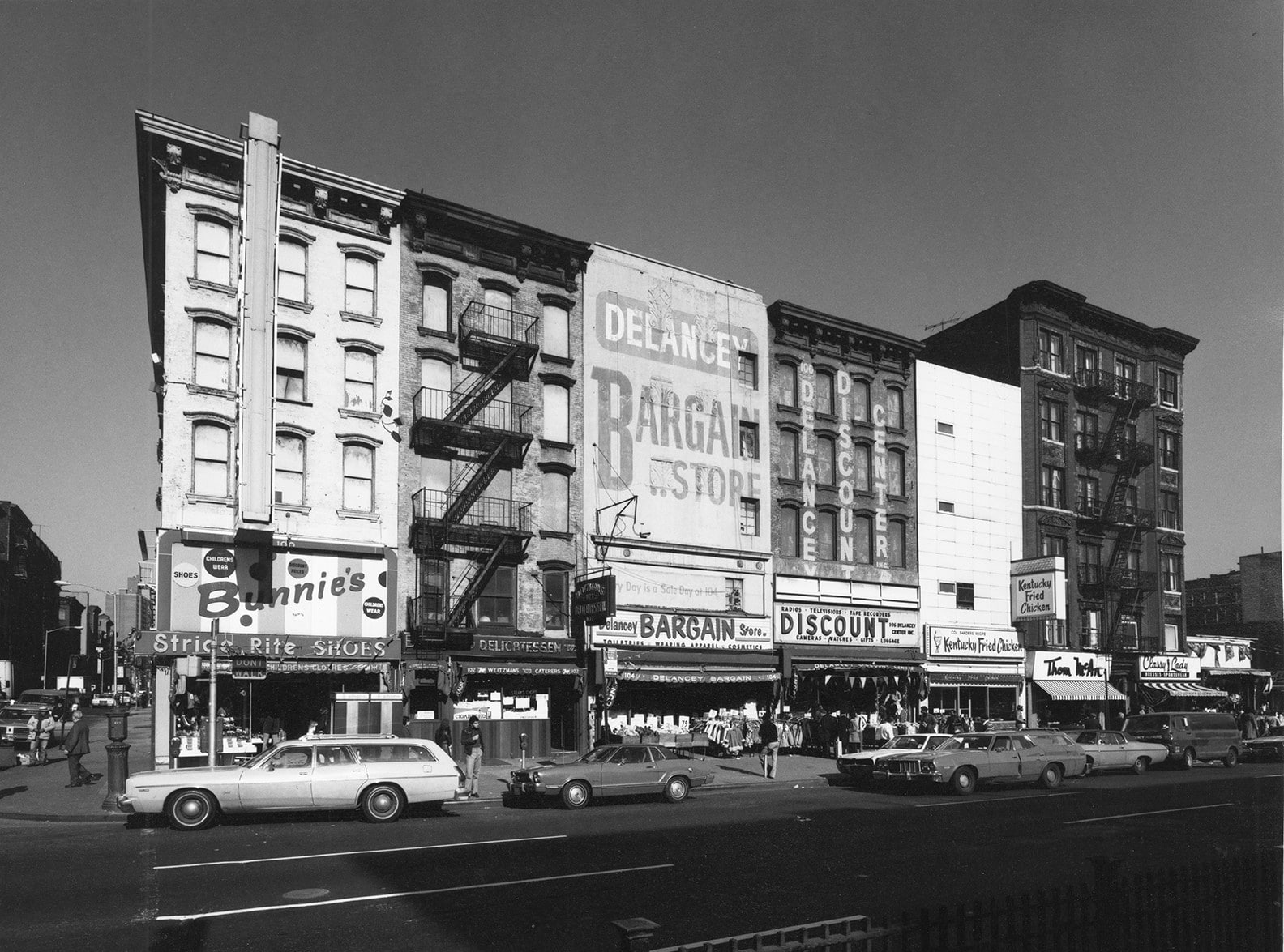 Cars pass in front of 6 neighboring tenements with street-level storefronts for "Delancey's Bargain Store", "Bunnie's Shoes", and "KFC"