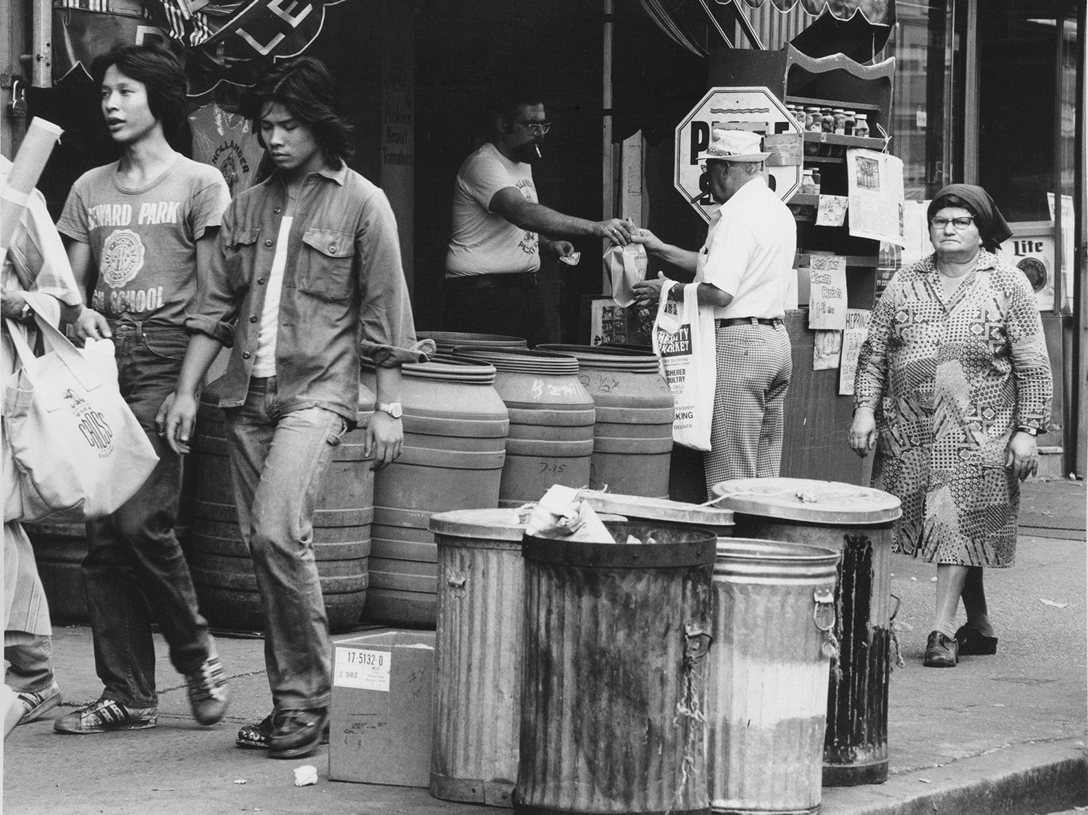 Two young adults walk casually past an older adult picking up a purchase from a shopkeeper on a street lined with bins and barrels