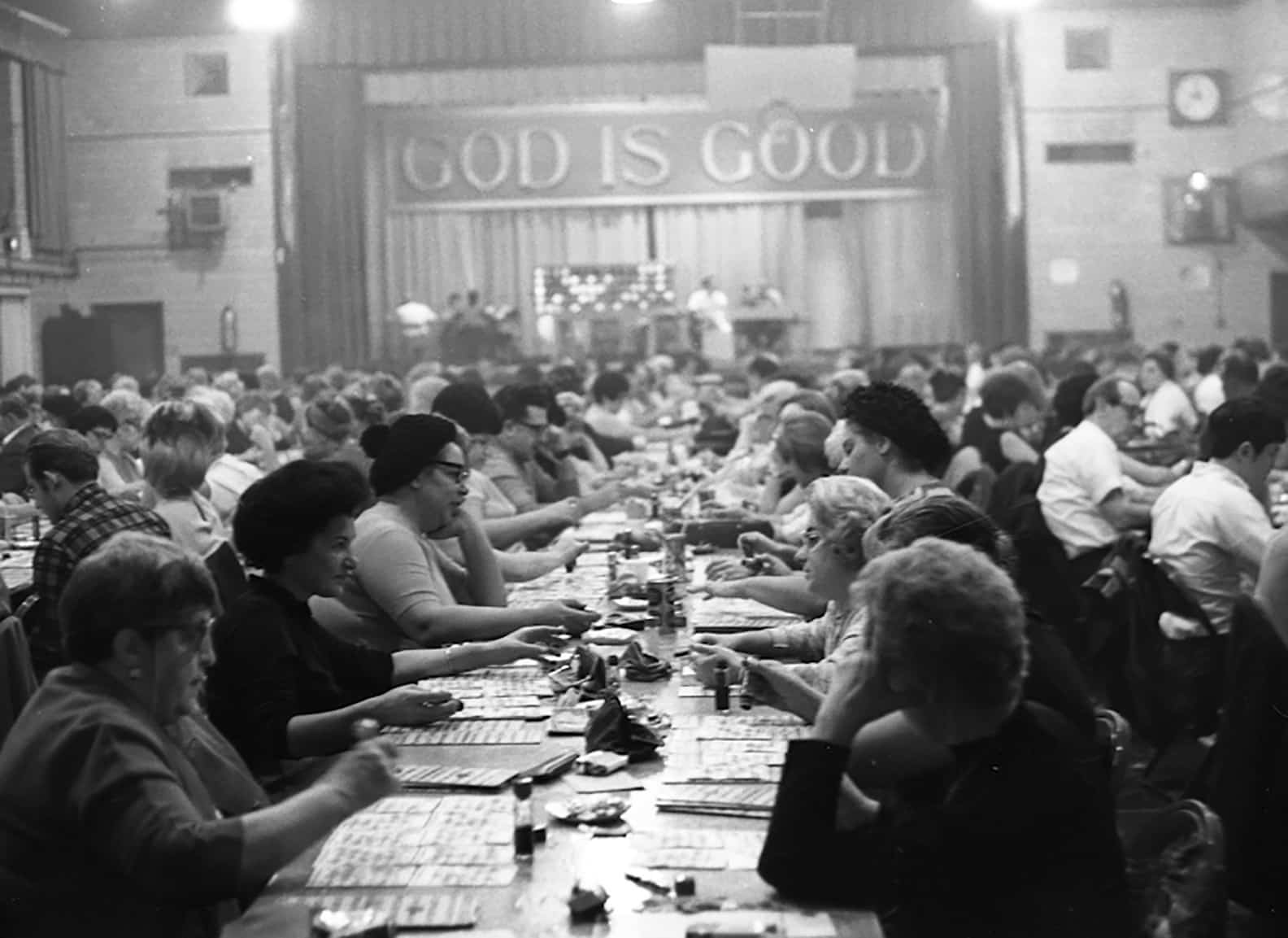 Rows of adults sitting at tables gathered in a large auditorium space playing bingo. A large sign reading "God is Good" hangs above the stage.