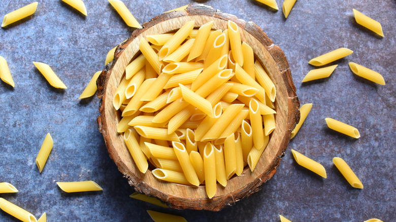 Wooden bowl of uncooked penne pasta on blue background