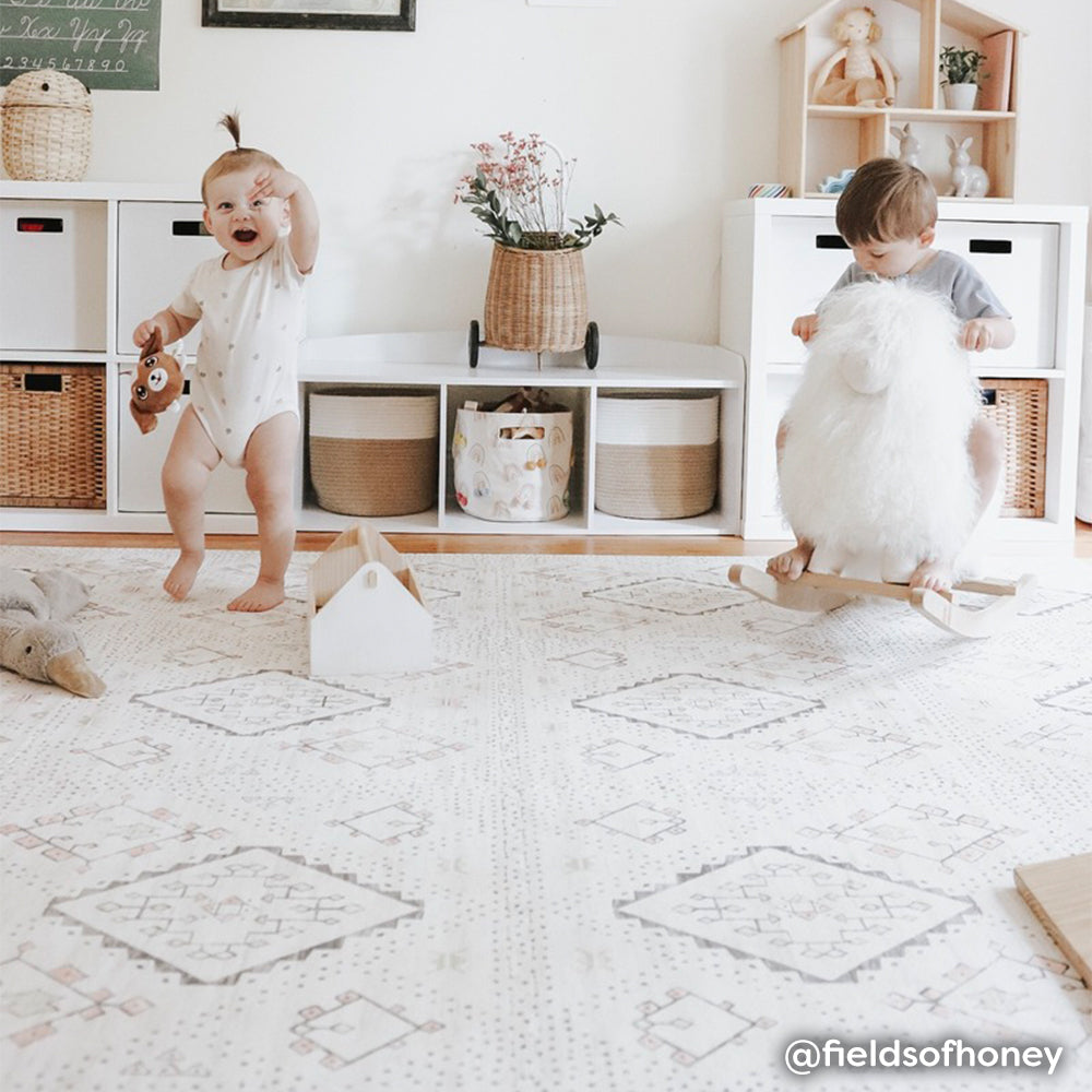 Ula Oat neutral boho print baby play mat shown in play room with two babies playing on play mat. @fieldsofhoney written in bottom right hand corner.