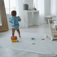 Ivory and white minimal geometric print baby play mat shown in living room with toys and baby walking on play mat. @kenzielucasking written in bottom right hand corner.