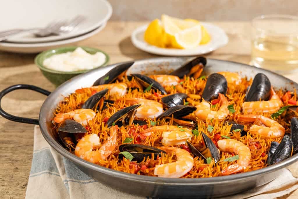 A close up of Catalan fideua in a skillet in front of a bowl of aioli, lemon wedges, a glass of wine, 2 plates and 2 forks.