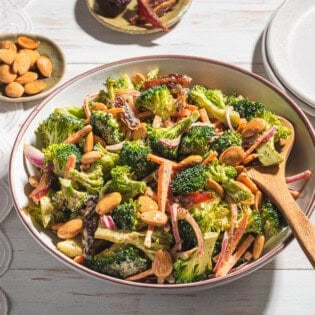 A photo of broccoli salad in a serving bowl with a wooden spoon. Next to this is a stack of 2 plates and small bowls of dates and toasted almonds.