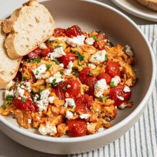 A close up of a serving of strapastada greek tomato scrambled eggs topped with crumbled feta and chopped parsley in a bowl with 2 slices of bread. Next to this is a cloth napkin and a plate of sliced bread.