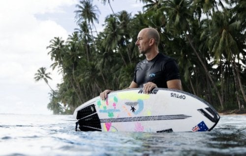 A surfer stands in the shallows with his surfboard floating in the water.