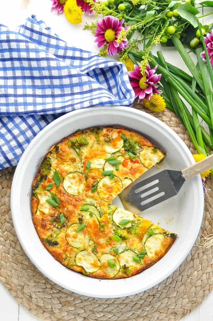 Overhead image of vegetable pie in round baking dish with a blue dish cloth and flowers beside it