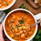 Overhead image of hands holding a white bowl full of creamy beef tomato soup with noodles on a wooden table.