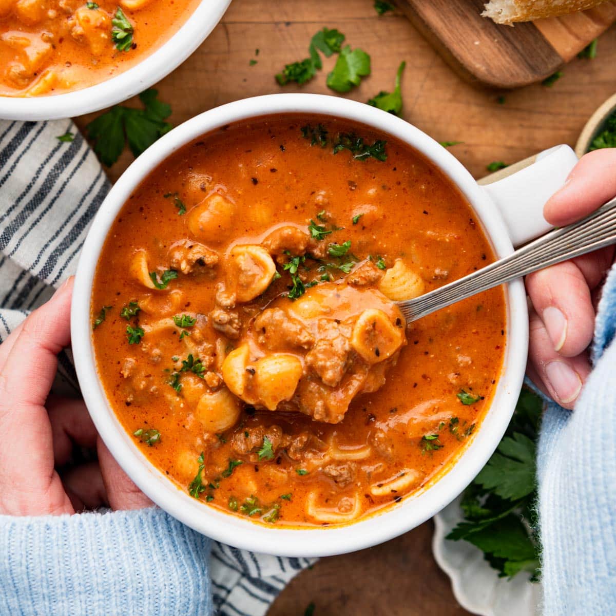 Square overhead shot of hands eating a bowl of creamy tomato soup with ground beef and noodles.