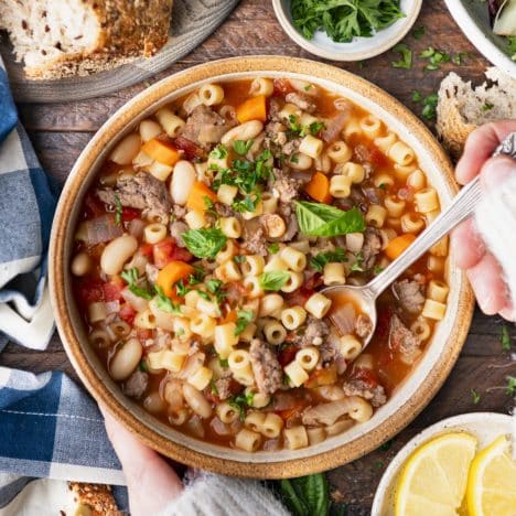 Square overhead shot of hands eating a bowl of pasta fagioli soup.