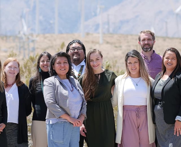 A multiracial and gender-diverse group of people working with Navajo Power, a fiscally sponsored partner of Tides, standing in front of wind turbines.