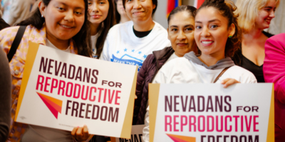 Group of women with dark hair. Two women hold signs that say Nevadans for Reproductive Freedom