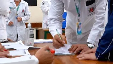 Imagem mostra médicos com jalecos brancos debruçados em uma mesa fazendo anotações.