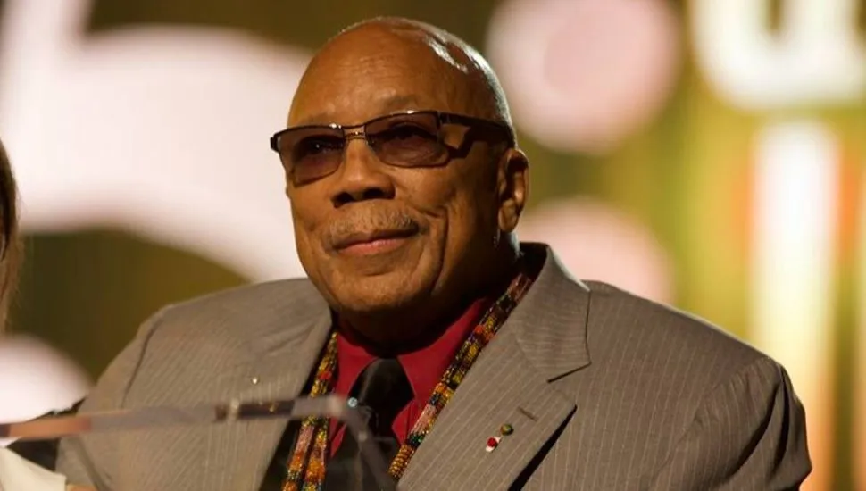 Imagem mostra Quincy Jones, com terno bege, camisa vermelha, óculos escuros e gravata preta.