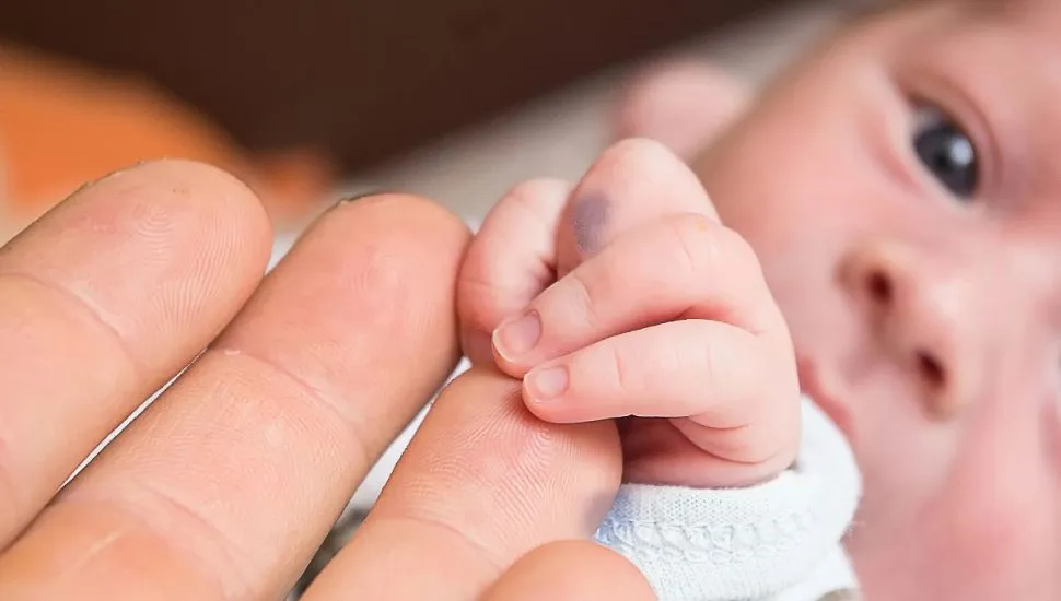 Na imagem, detalhe do rosto de bebê recém-nascido, que segura nos dedos de um adulto.